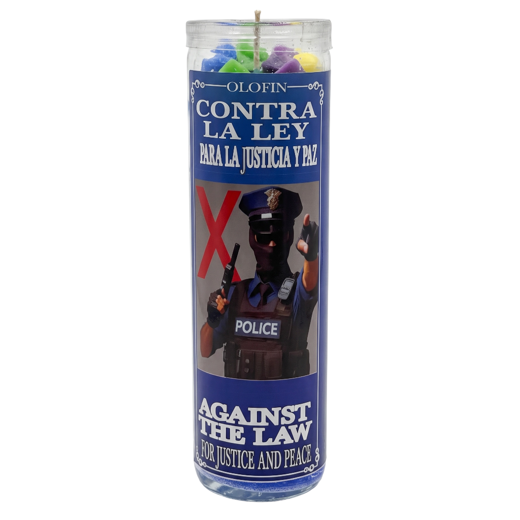 A tall glass prayer candle with a clear top and multicolored wax pieces visible inside. The candle has a blue printed label with Spanish and English text reading 'Olofin Contra La Ley Para La Justicia Y Paz' and 'Against The Law For Justice And Peace,' plus an image of a police officer and a large red X.