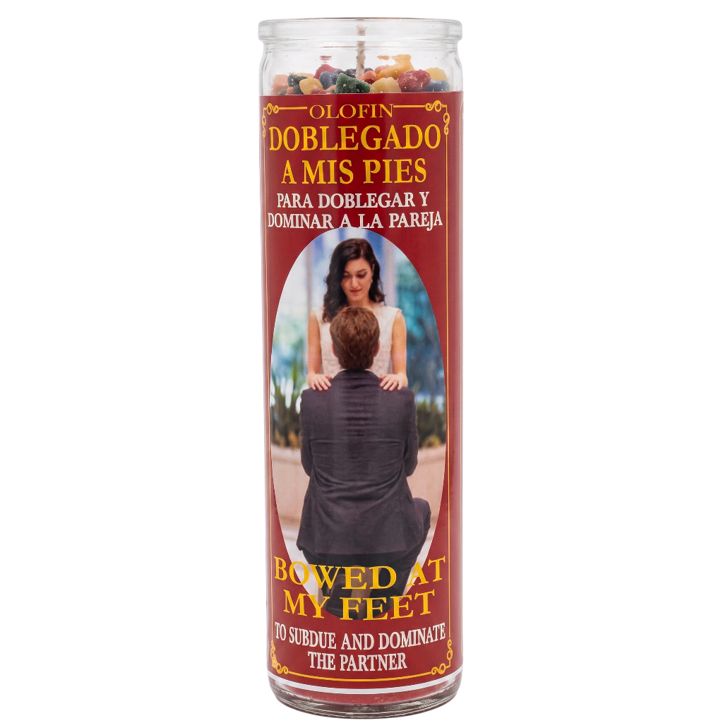 A tall glass jar candle with a red label featuring Spanish and English text, an oval photo of a couple, and multicolored beads visible at the top. The label includes the phrase 'BOWED AT MY FEET'.